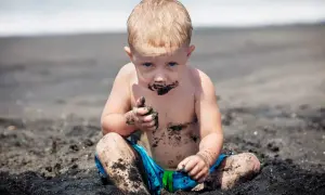 Petit garçon blond, couvert de sable noir, semble manger de l'argile sur la plage. Il a les mains pleines de terre.