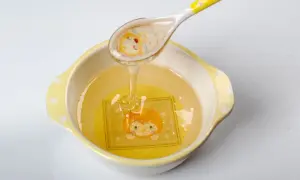 Une cuillère verse du miel doré dans un bol orné d'un dessin d'animal. Ce miel, d'un âge minimum, coule en un fin filet.