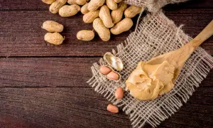 Une cuillère de beurre de cacahuète crémeux, avec des cacahuètes entières et décortiquées à proximité. Attention, la cacahuète peut présenter un danger pour les personnes allergiques.