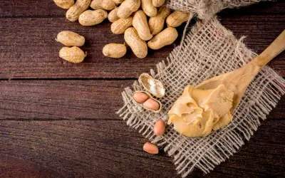 Une cuillère de beurre de cacahuète crémeux, avec des cacahuètes entières et décortiquées à proximité. Attention, la cacahuète peut présenter un danger pour les personnes allergiques.
