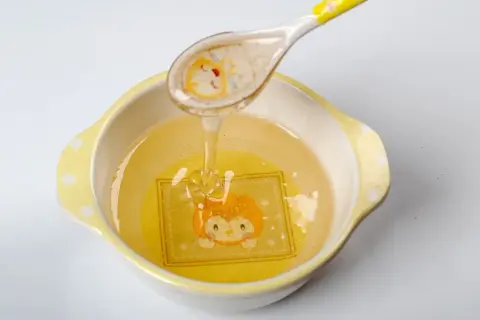 Une cuillère verse du miel doré dans un bol orné d'un dessin d'animal. Ce miel, d'un âge minimum, coule en un fin filet.