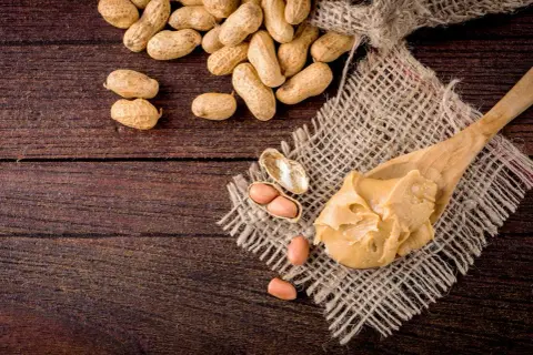 Une cuillère de beurre de cacahuète crémeux, avec des cacahuètes entières et décortiquées à proximité. Attention, la cacahuète peut présenter un danger pour les personnes allergiques.