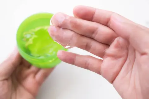 Main tenant une main, une autre prélève un gel transparent d'un pot vert. Attention à l'allergie à l'aloe vera.