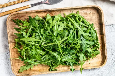 Un plateau en bois déborde de roquette salade fraîche, prête à être dégustée avec des couverts en bois et métal.