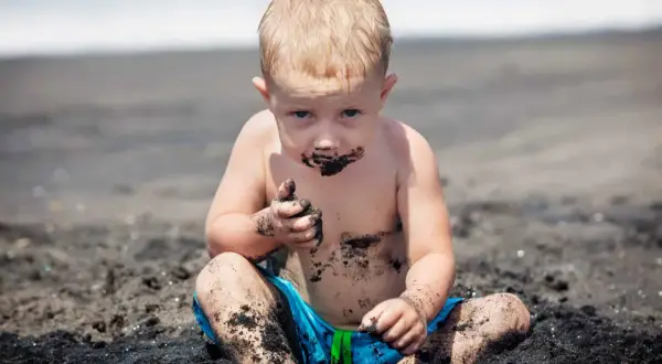 Petit garçon blond, couvert de sable noir, semble manger de l'argile sur la plage. Il a les mains pleines de terre.