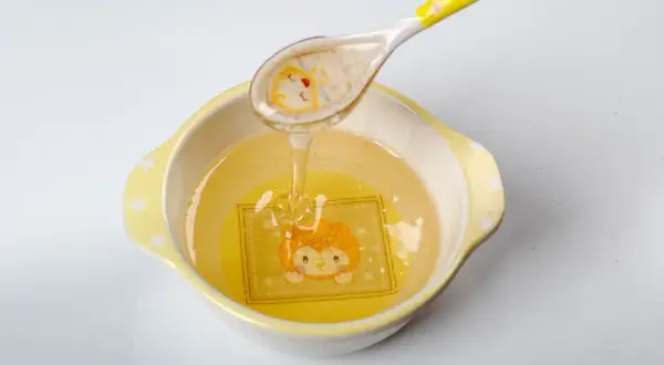 Une cuillère verse du miel doré dans un bol orné d'un dessin d'animal. Ce miel, d'un âge minimum, coule en un fin filet.