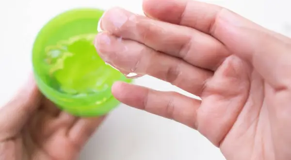 Main tenant une main, une autre prélève un gel transparent d'un pot vert. Attention à l'allergie à l'aloe vera.