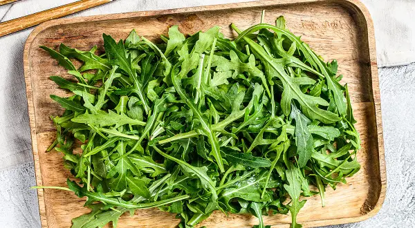 Un plateau en bois déborde de roquette salade fraîche, prête à être dégustée avec des couverts en bois et métal.
