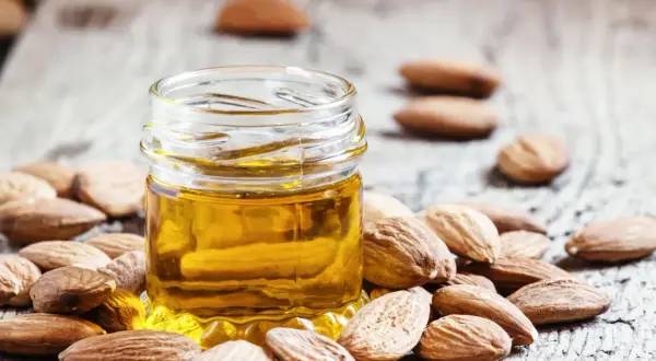 Un petit pot d'huile d'amande douce, entouré d'amandes entières sur fond de bois. Découvrez les bienfaits de l'huile d'amande douce pour la peau.