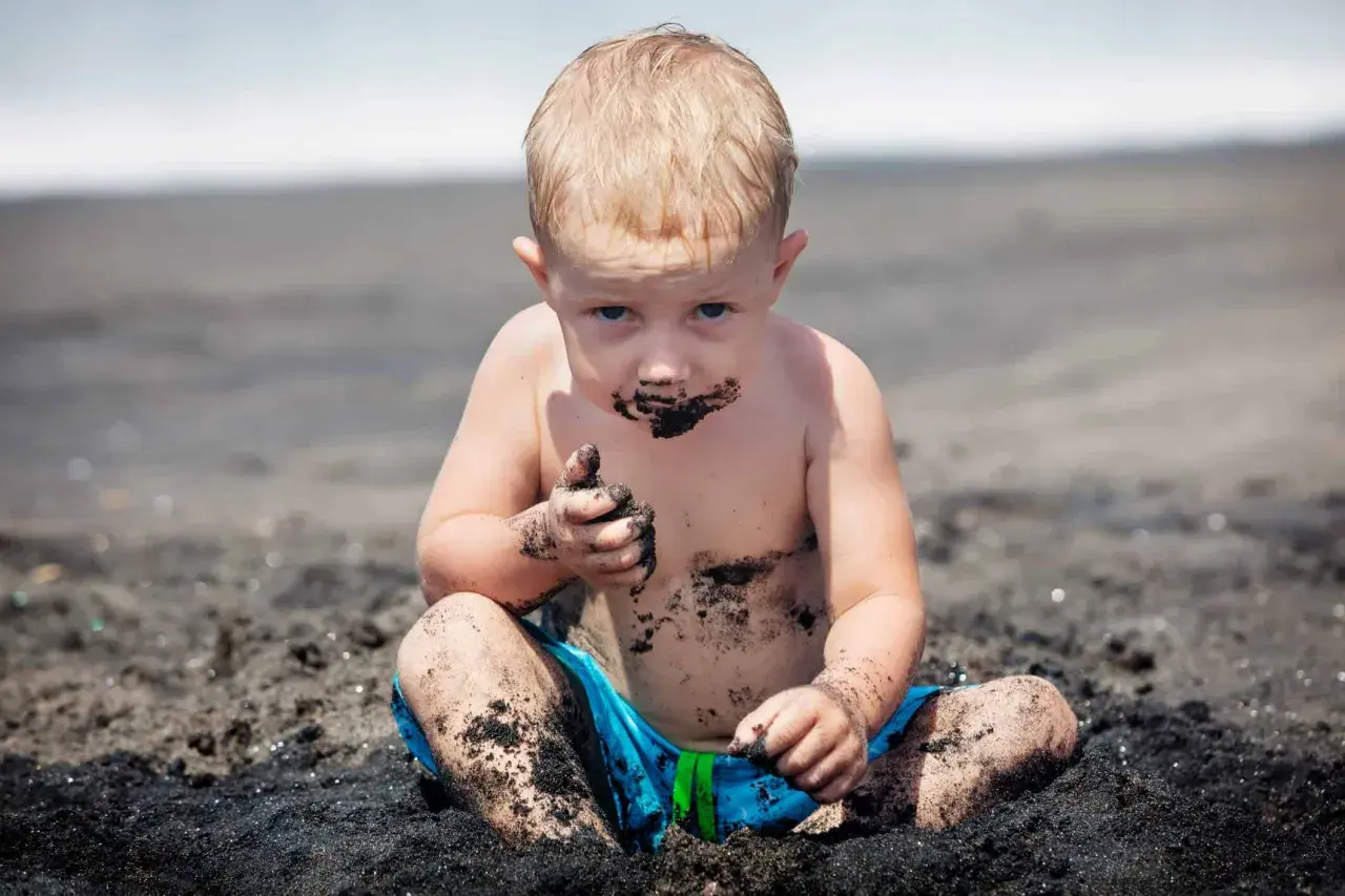 Petit garçon blond, couvert de sable noir, semble manger de l'argile sur la plage. Il a les mains pleines de terre.