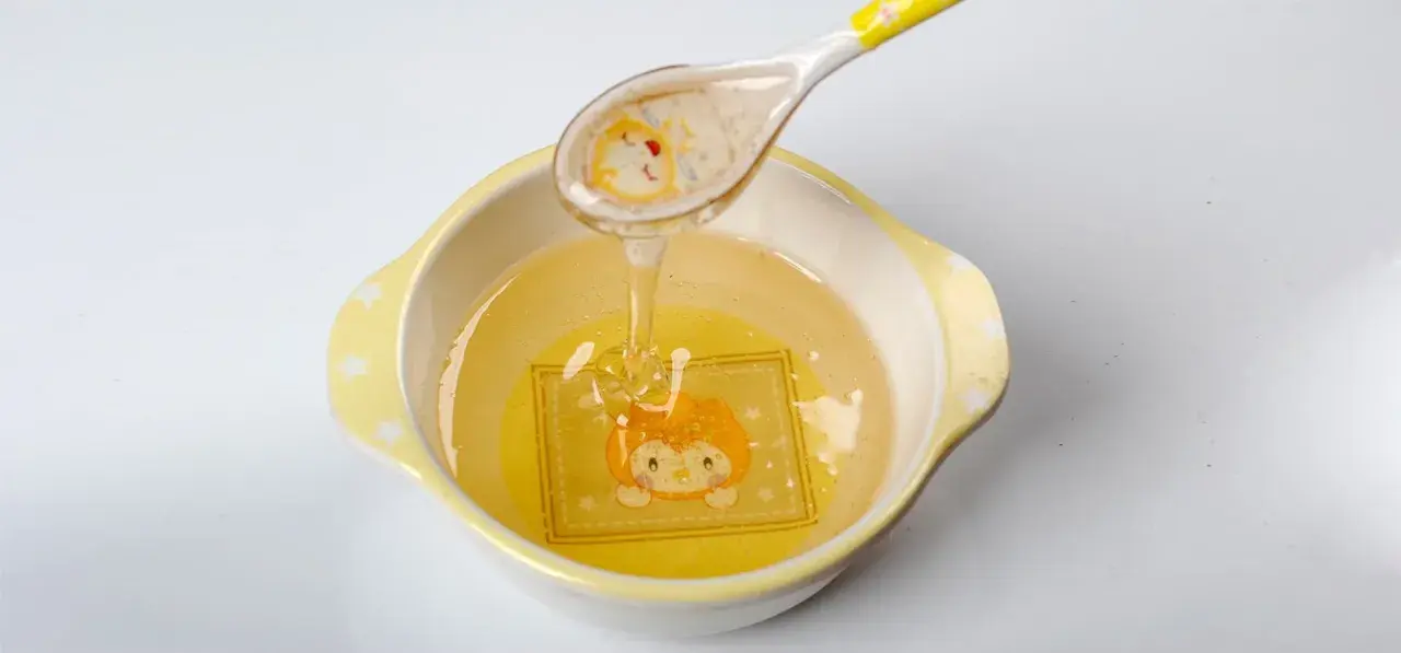 Une cuillère verse du miel doré dans un bol orné d'un dessin d'animal. Ce miel, d'un âge minimum, coule en un fin filet.