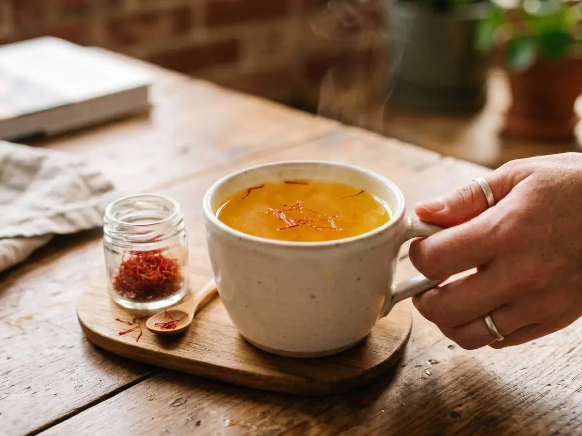 Une tasse de boisson dorée fumante, infusée de safran dose maximale. À côté, un petit pot de pistils rouges et une cuillère en bois.