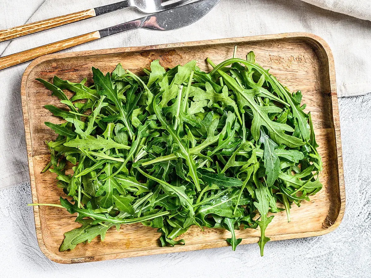 Un plateau en bois déborde de roquette salade fraîche, prête à être dégustée avec des couverts en bois et métal.