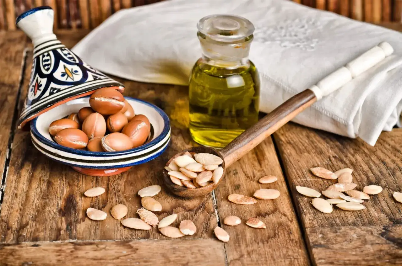 Huile d'argan bienfaits : amandes d'argan dans un plat traditionnel, une cuill&egrave;re remplie d'amandes concass&eacute;es et une petite fiole d'huile dor&eacute;e sur fond de bois.
