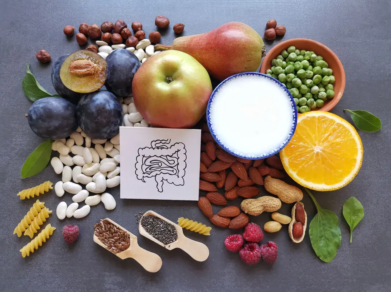 Assortiment de fruits, légumes, noix et graines, dont des prunes, une pomme, une poire, des haricots, des amandes, des cacahuètes, des framboises, des pois, des graines de lin, des graines de chia, des pâtes et une tranche d'orange. Ces 13 aliments qui...