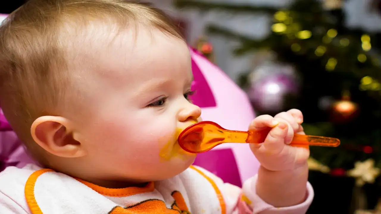 B&eacute;b&eacute; mangeant avec une cuill&egrave;re orange, un bavoir autour du cou. Un sapin de No&euml;l d&eacute;cor&eacute; est visible en arri&egrave;re-plan. Le miel est interdit avant l'&acirc;ge minimum d'un an.