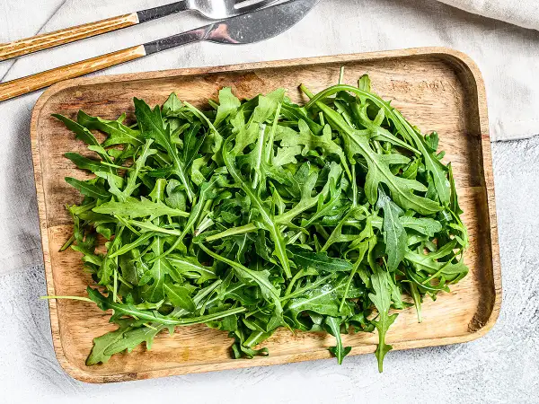 Un plateau en bois déborde de roquette salade fraîche, prête à être dégustée avec des couverts en bois et métal.